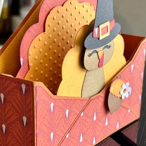 May include: A handmade Thanksgiving card with a turkey in a pilgrim hat. The card is made with layers of orange, yellow, and brown cardstock. It is decorated with a small flower and a leaf, and has a textured finish.