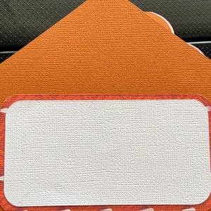 May include: An orange envelope with a textured surface and a rectangular label with rounded corners. The label has a white centre and a decorative border with small white accents.