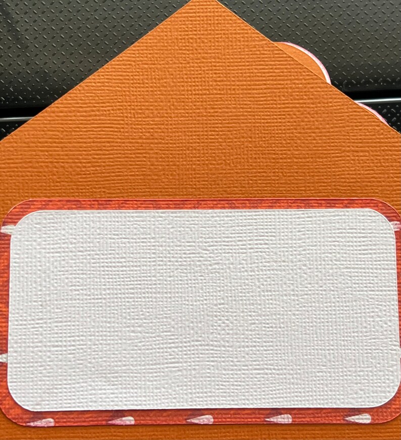 May include: An orange envelope with a textured surface and a rectangular label with rounded corners. The label has a white centre and a decorative border with small white accents.