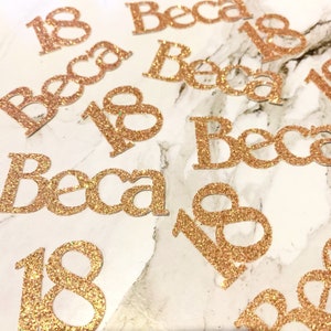 May include: Gold glitter confetti with the name "Becca" and the number "18" on a marble background.