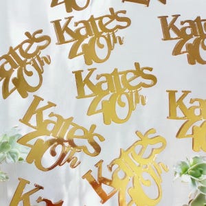 Könnte beinhalten: Goldfarbene, reflektierende Tortenaufsätze mit dem Text "Kate's 40th" in einer eleganten Schrift. Die Aufsätze sind auf einem weißen Hintergrund verstreut, was auf ein festliches Ereignis hindeutet. Geeignet für einen runden Geburtstag.