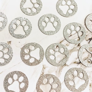 May include: Silver glitter paw print confetti circles. The circles are about 1 inch in diameter and have a silver glitter finish. The paw prints are cut out of the center of the circles.