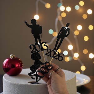 Könnte beinhalten: Ein schwarzer Glitzer-Kuchenaufsatz mit Silhouetten eines Babys, eines Kindes mit Rucksack und eines Mannes im Anzug, mit den Jahren 2008, 2013 und 2026. Eine rote Weihnachtskugel und eine weiße Torte im Hintergrund.