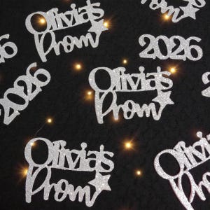 Può includere: Ritagli argentati glitterati con la scritta "Olivia's Prom" e il numero "2026" sparsi su una superficie nera. I ritagli sono illuminati da piccole luci di fata dai toni caldi, creando un'atmosfera festosa.