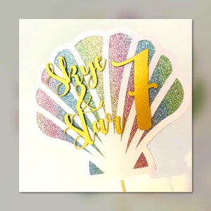 Op de afbeelding: Een witte glitter schelp taarttopper met een gouden glitter "Skye & Starr" en het nummer "1" in gouden glitter.