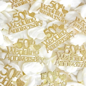 Pode incluir: Toppers de bolo dourados com glitter "50º Aniversário de Casamento" com detalhes de coração. Os toppers estão espalhados sobre uma cama de pétalas de flores brancas. O texto está em fonte cursiva.