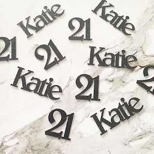 May include: Black glitter confetti with the name "Katie" and the number "21" scattered on a white marble surface.