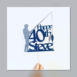 May include: A blue glitter cake topper with a silhouette of a person fishing. The topper says "Happy 40th Steve" and has two fish on the line.