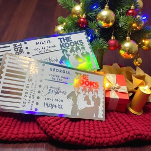 May include: Two concert tickets for "The Kooks" with holographic details, featuring the band's image and the names "Millie" and "Georgia." The tickets are presented with a red gift box, a lit candle, and a Christmas tree with baubles.