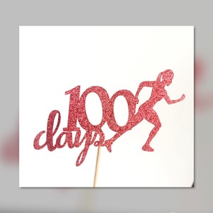 May include: A pink glitter cake topper with the number 100 and the word "days" in cursive. A pink glitter silhouette of a runner is next to the number 100.