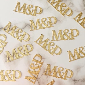 May include: Gold glitter cutouts of the letters "M&D" are scattered across a white and gray marble surface. The letters are in a serif font and are slightly angled. The overall theme is wedding or event decor.