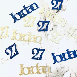 May include: Gold and blue glitter confetti with the name "Jordan" and the number "27" scattered on a white surface.