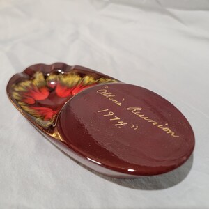 May include: A vintage ashtray with a deep red and gold floral design. The ashtray has a built-in rest and a separate oval section with the inscription "Allen's Reunion 1974."