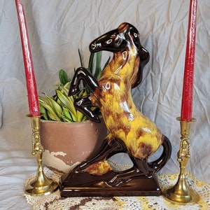 May include: A ceramic horse figurine with brown and yellow glaze. The horse is rearing up on its hind legs. The figurine is on a white crocheted doily with two brass candle holders and red candles.