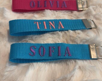 May include: Three fabric wristlet keychains with silver-tone metal hardware. The top strap is hot pink with the name OLIVIA embroidered in purple. The middle strap is blue with TINA in orange. The bottom strap is blue with SOFIA in purple.
