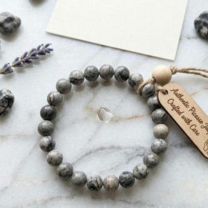 May include: A beaded bracelet made of gray Picasso jasper stones. The bracelet is accompanied by a wooden tag that reads "Authentic Picasso Jasper - Crafted with Care." The bracelet is displayed with decorative stones and lavender sprigs.