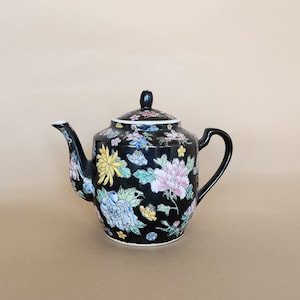 Peut inclure: Une théière noire avec un motif floral dans des tons de rose, jaune, bleu et vert. La théière a un couvercle et une poignée.