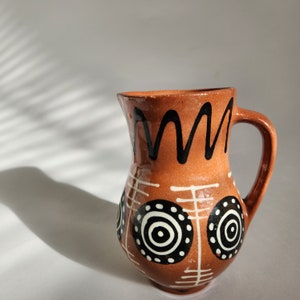 May include: A terracotta ceramic jug with a handle, decorated with black and white geometric patterns. The jug has a warm, earthy tone and is likely used for serving drinks. The design includes circles and wavy lines.