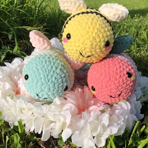 Puede incluir: Tres animales de peluche tejidos a crochet: una abeja amarilla y negra, un animal rayado azul claro y blanco y un animal rayado rosa y blanco. Los animales est&aacute;n sentados en un arreglo de flores blancas.