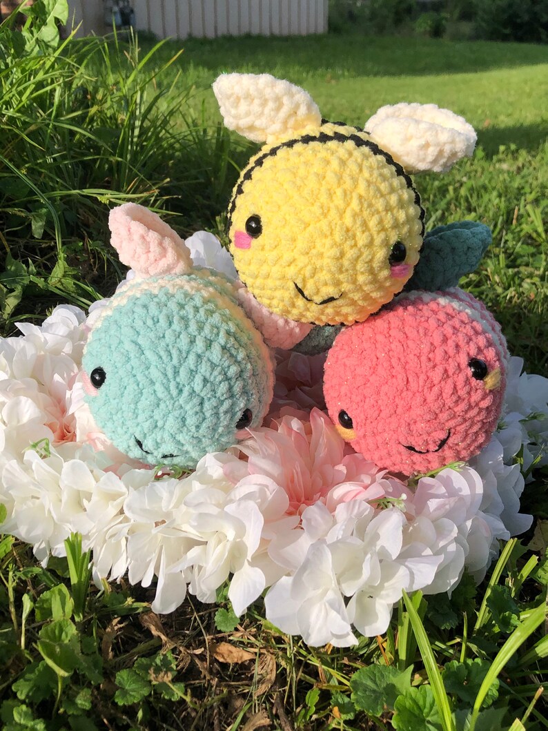 Puede incluir: Tres animales de peluche tejidos a crochet: una abeja amarilla y negra, un animal rayado azul claro y blanco y un animal rayado rosa y blanco. Los animales est&aacute;n sentados en un arreglo de flores blancas.