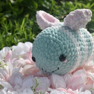 Puede incluir: Un juguete de peluche de abeja tejido a crochet con un cuerpo a rayas azul claro y blanco, orejas rosas y ojos negros. La abeja est&aacute; sentada en una cama de p&eacute;talos de flores blancas y rosas.
