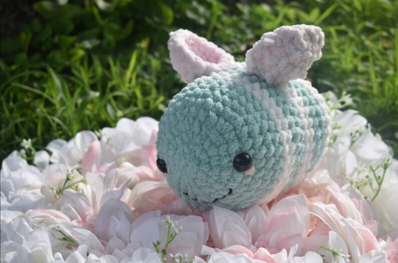 Puede incluir: Un juguete de peluche de abeja tejido a crochet con un cuerpo a rayas azul claro y blanco, orejas rosas y ojos negros. La abeja est&aacute; sentada en una cama de p&eacute;talos de flores blancas y rosas.