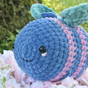 Puede incluir: Un juguete de peluche de crochet con forma de abeja con rayas azules y rosas y alas de color verde azulado. La abeja tiene ojos negros y una sonrisa cosida.