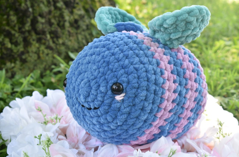 Puede incluir: Un juguete de peluche de crochet con forma de abeja con rayas azules y rosas y alas de color verde azulado. La abeja tiene ojos negros y una sonrisa cosida.