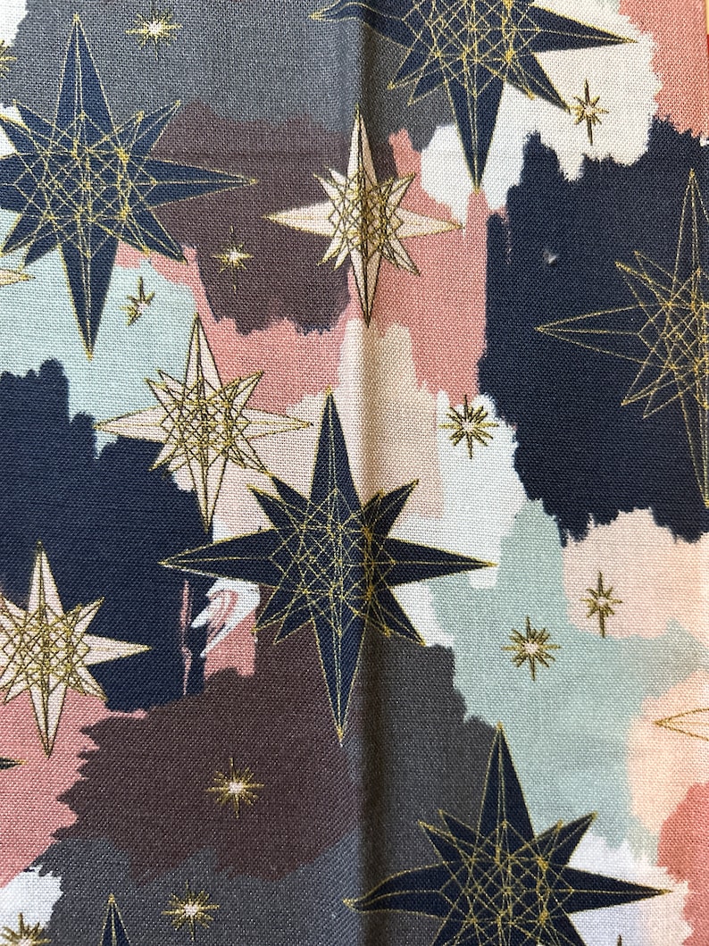 May include: A fabric with a repeating pattern of gold stars on a background of pink, brown, and blue watercolor-like shapes.