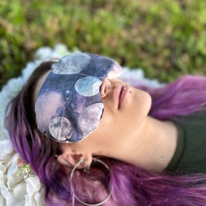 May include: A person with purple hair is wearing a sleep mask with a moon and stars print. The mask is covering their eyes and they are lying down on a white surface.