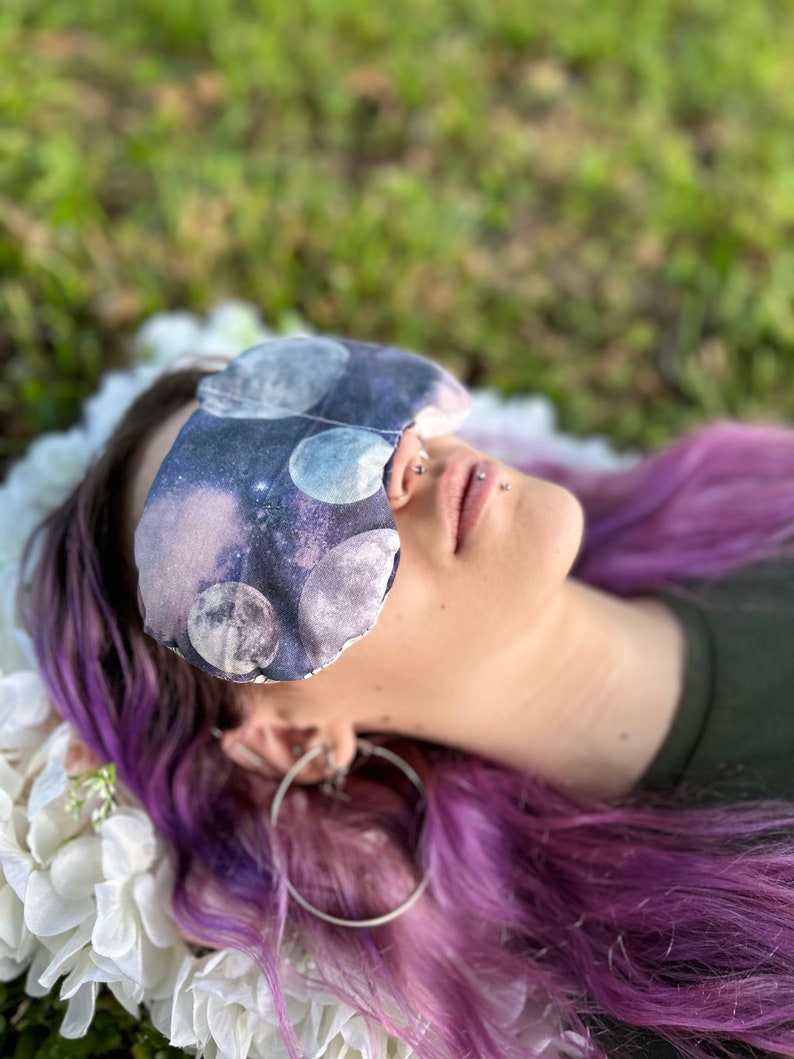 May include: A person with purple hair is wearing a sleep mask with a moon and stars print. The mask is covering their eyes and they are lying down on a white surface.