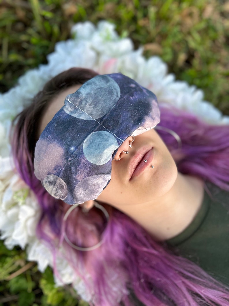 May include: A person with purple hair wearing a sleep mask with a galaxy print. The mask is made of soft fabric and has a comfortable fit.