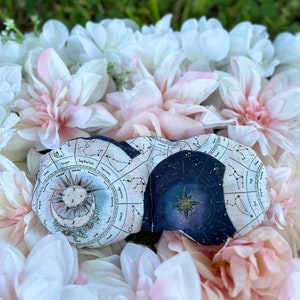 May include: A sleep mask with a celestial design featuring a sun, moon, and stars against a navy blue background. The mask is surrounded by white and pink flowers.