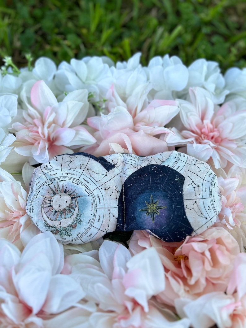 May include: A sleep mask with a celestial design featuring a sun, moon, and stars against a navy blue background. The mask is surrounded by white and pink flowers.