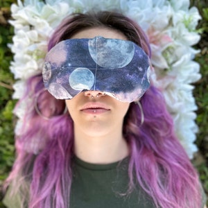 May include: A person with purple hair wearing a sleep mask with a galaxy print. The sleep mask has a dark blue background with white and gray moons and stars.