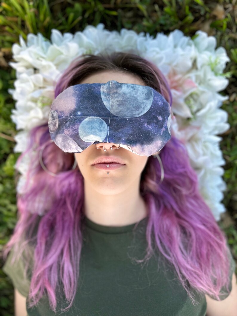 May include: A person with purple hair wearing a sleep mask with a galaxy print. The sleep mask has a dark blue background with white and gray moons and stars.