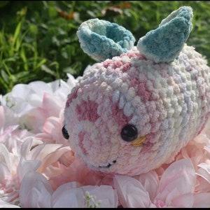 Puede incluir: Un animal de peluche tejido a crochet con forma de abeja, hecho con hilo rosa, blanco y azul. La abeja tiene alas grandes de color azul y ojos negros con detalles amarillos.