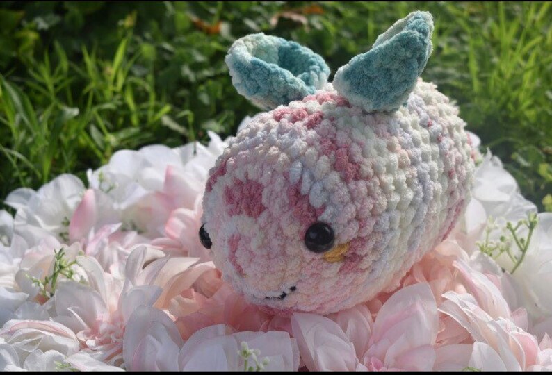 Puede incluir: Un animal de peluche tejido a crochet con forma de abeja, hecho con hilo rosa, blanco y azul. La abeja tiene alas grandes de color azul y ojos negros con detalles amarillos.