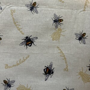 May include: A white fabric with a repeating pattern of bumblebees and the text "Bumblebee" in a script font. The bumblebees are black and yellow with brown wings.