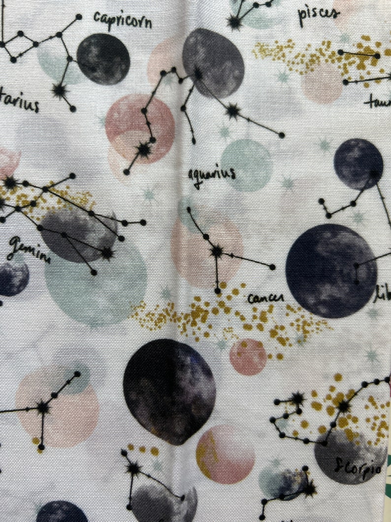 May include: White fabric with a repeating pattern of constellations and planets. The constellations are outlined in black and the planets are painted in shades of gray and pink. The constellations are labeled with their astrological signs: Capricorn, Aquarius, Pisces, Taurus, Gemini, Cancer, Libra, Scorpio.