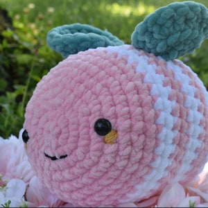 Puede incluir: Un juguete de peluche de crochet rosa y blanco con forma de abeja con alas de fieltro verde. La abeja tiene ojos negros con detalles amarillos y una boca sonriente.