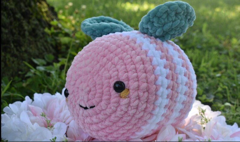 Puede incluir: Un juguete de peluche de crochet rosa y blanco con forma de abeja con alas de fieltro verde. La abeja tiene ojos negros con detalles amarillos y una boca sonriente.