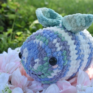Puede incluir: Un animal de peluche tejido a crochet con forma de abeja, hecho con hilo azul, blanco, verde y morado. La abeja tiene grandes ojos negros y una peque&ntilde;a boca rosa.