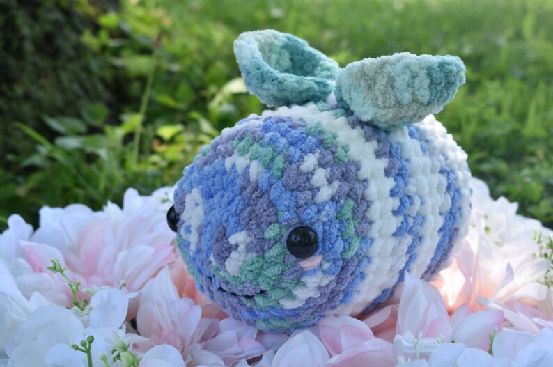 Puede incluir: Un animal de peluche tejido a crochet con forma de abeja, hecho con hilo azul, blanco, verde y morado. La abeja tiene grandes ojos negros y una peque&ntilde;a boca rosa.