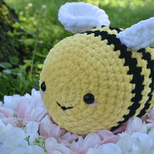 Puede incluir: Un juguete de peluche de abeja tejido a crochet con rayas amarillas y negras, alas blancas y una cara sonriente. La abeja est&aacute; sentada en una cama de flores blancas y rosas.