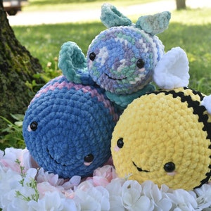 Puede incluir: Tres juguetes de peluche de crochet: una ballena azul y rosa, un pez verde y morado y una abeja amarilla y negra. Los juguetes est&aacute;n sentados en un lecho de flores blancas.