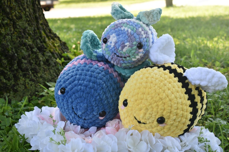 Puede incluir: Tres juguetes de peluche de crochet: una ballena azul y rosa, un pez verde y morado y una abeja amarilla y negra. Los juguetes est&aacute;n sentados en un lecho de flores blancas.