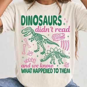 Puede incluir: Una camiseta de manga corta color crema con un estampado gráfico en verde y rosa. El texto dice DINOSAURS didn't read and we know WHAT HAPPENED TO THEM, acompañado de ilustraciones de esqueletos de dinosaurios y varios huesos fósiles.