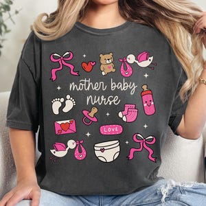 Puede incluir: Camiseta gris oscuro con un diseño gráfico que presenta ilustraciones relacionadas con bebés en rosa y blanco. El texto "mother baby nurse" está centrado en la camiseta. El diseño incluye lazos, un osito de peluche, un chupete y pies de bebé.