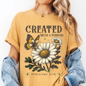 以下が含まれることがあります： マスタードイエローのTシャツに、蝶とデイジーのヴィンテージスタイルのグラフィックがプリントされています。Tシャツには「CREATED WITH A PURPOSE」と「EPHESIANS 2:10」の文字がプリントされています。